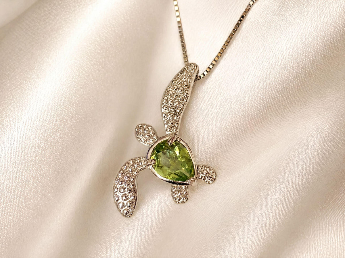 A silver necklace with a pendant featuring a green gemstone, resembling a turtle or sea turtle, is displayed against a white background.