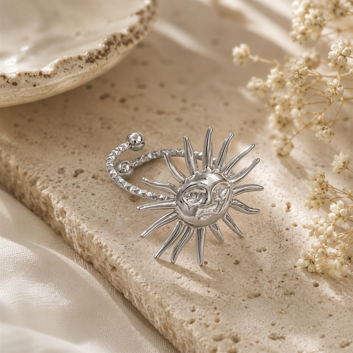 A silver ring with a sun-shaped design is placed on a beige surface, surrounded by small white flowers.
