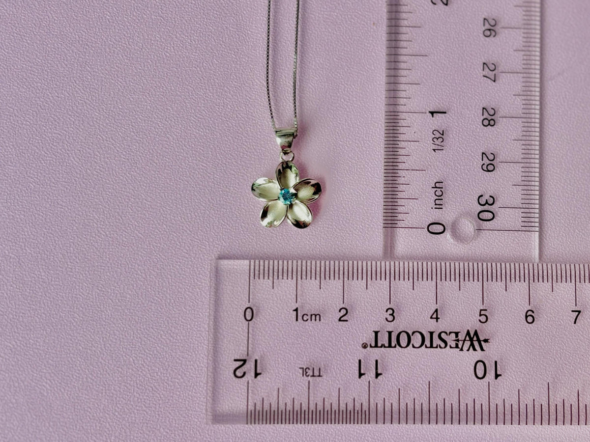 A silver flower pendant necklace is placed on a pink background, with a clear ruler for scale placed next to it.