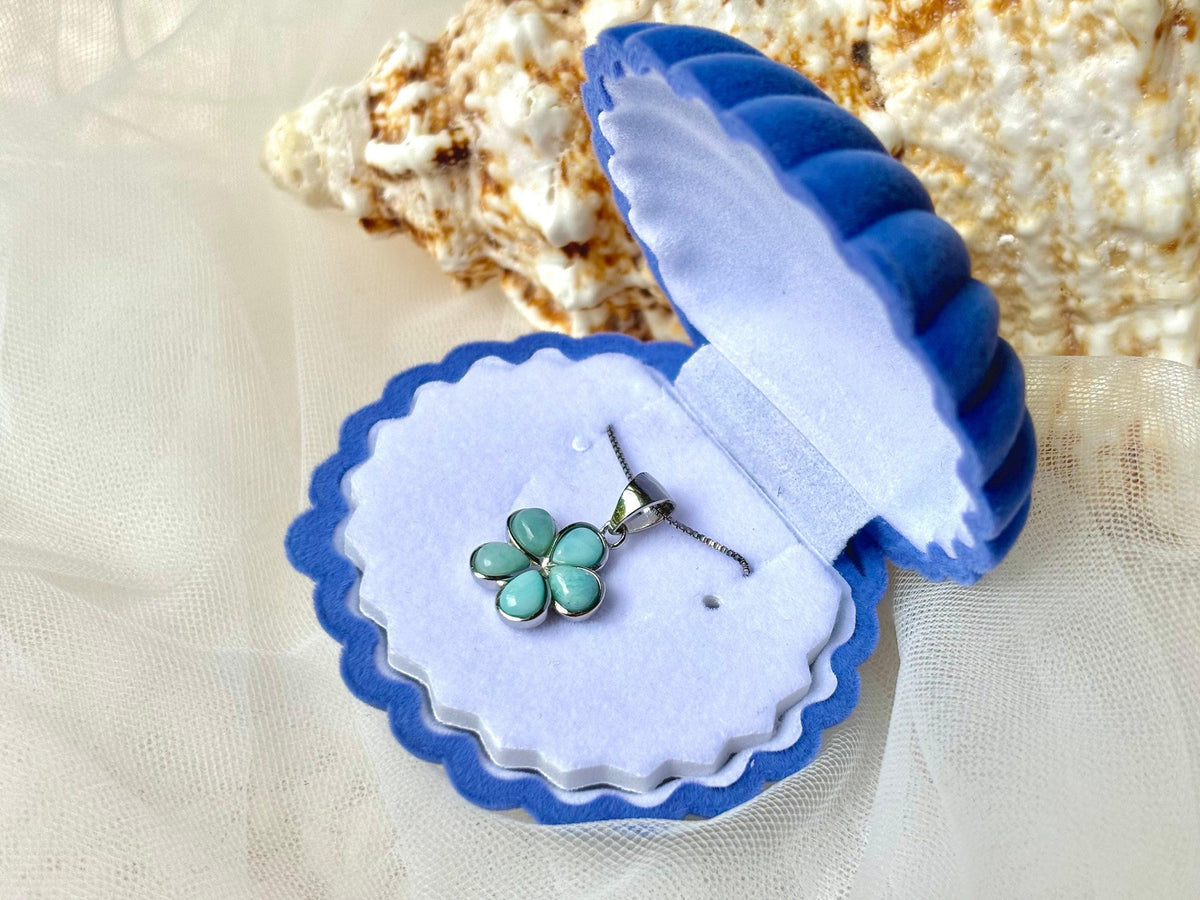A necklace with a blue pendant featuring a flower design is displayed in a blue and white scalloped box, placed on a white background.