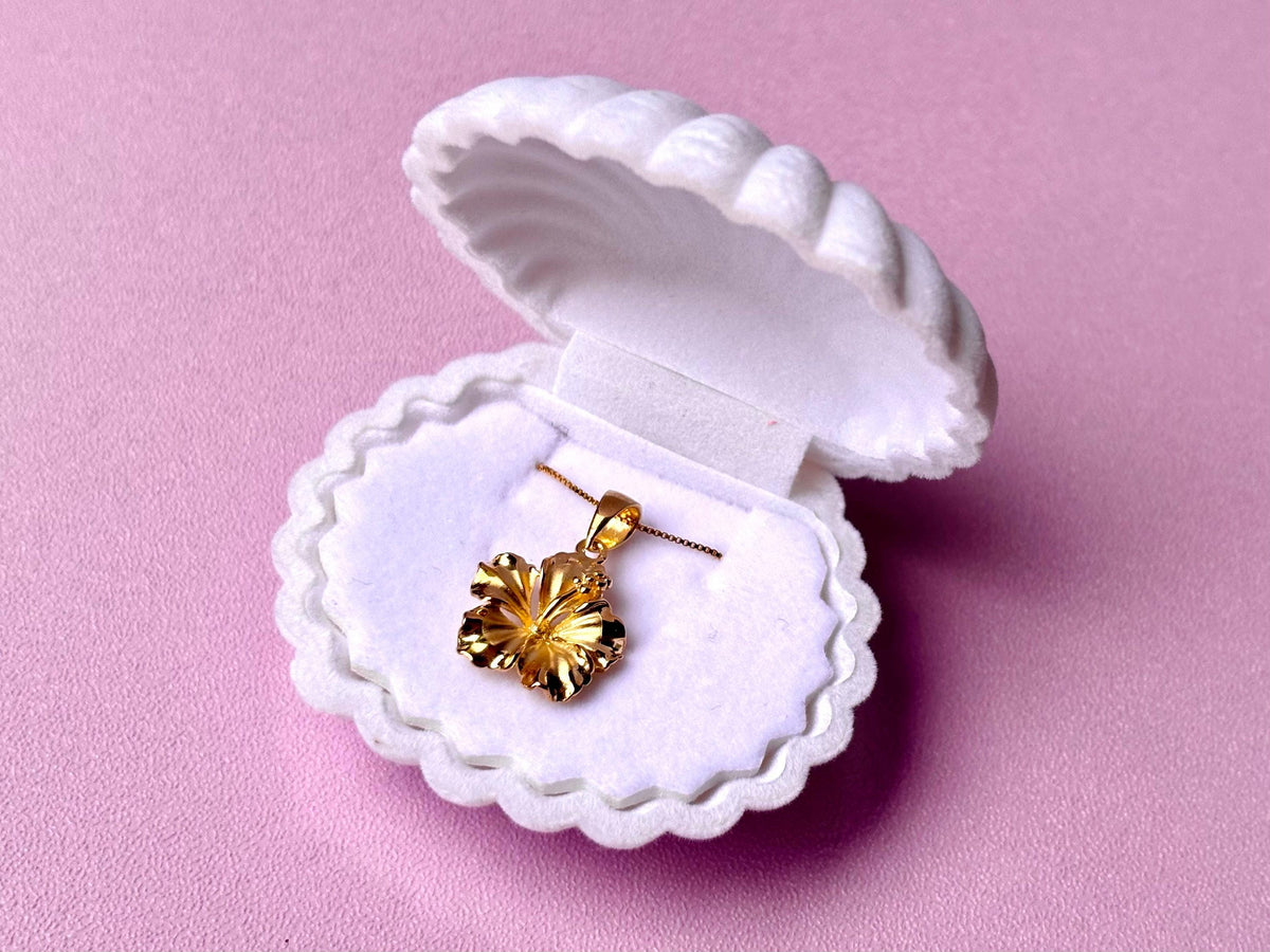 A gold necklace pendant is displayed in a white, scalloped-edge jewelry box against a pink background.