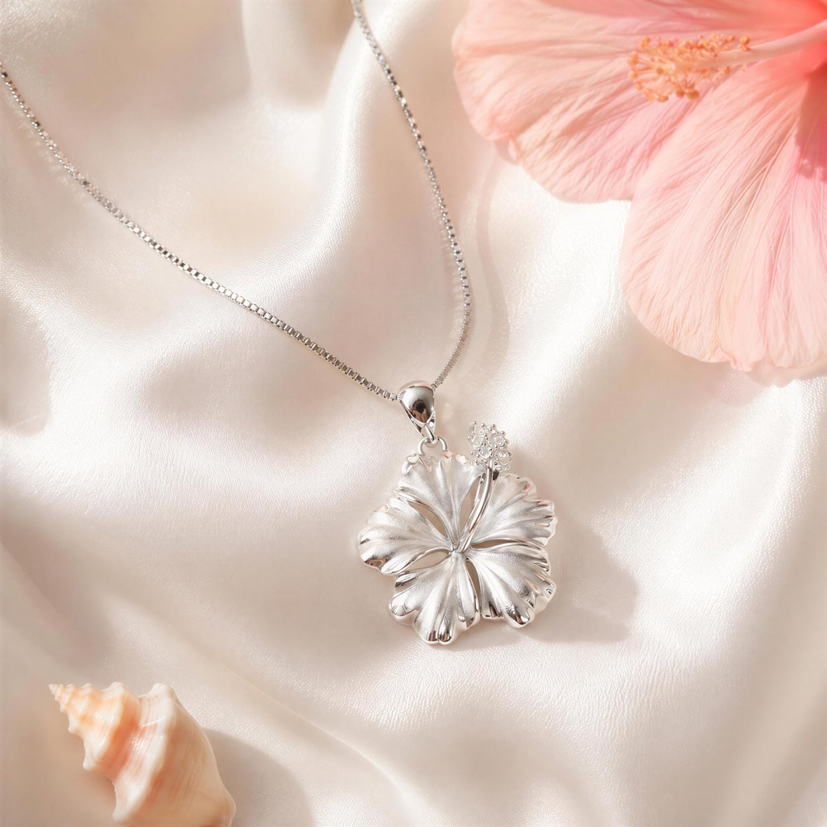 A delicate silver necklace with a pendant featuring a large, intricately designed flower rests on a soft, white fabric surface.