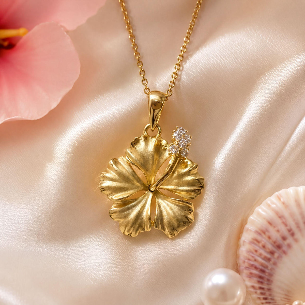 A gold necklace with a large, intricately designed flower pendant is displayed against a soft pink background, accompanied by a few seashells.