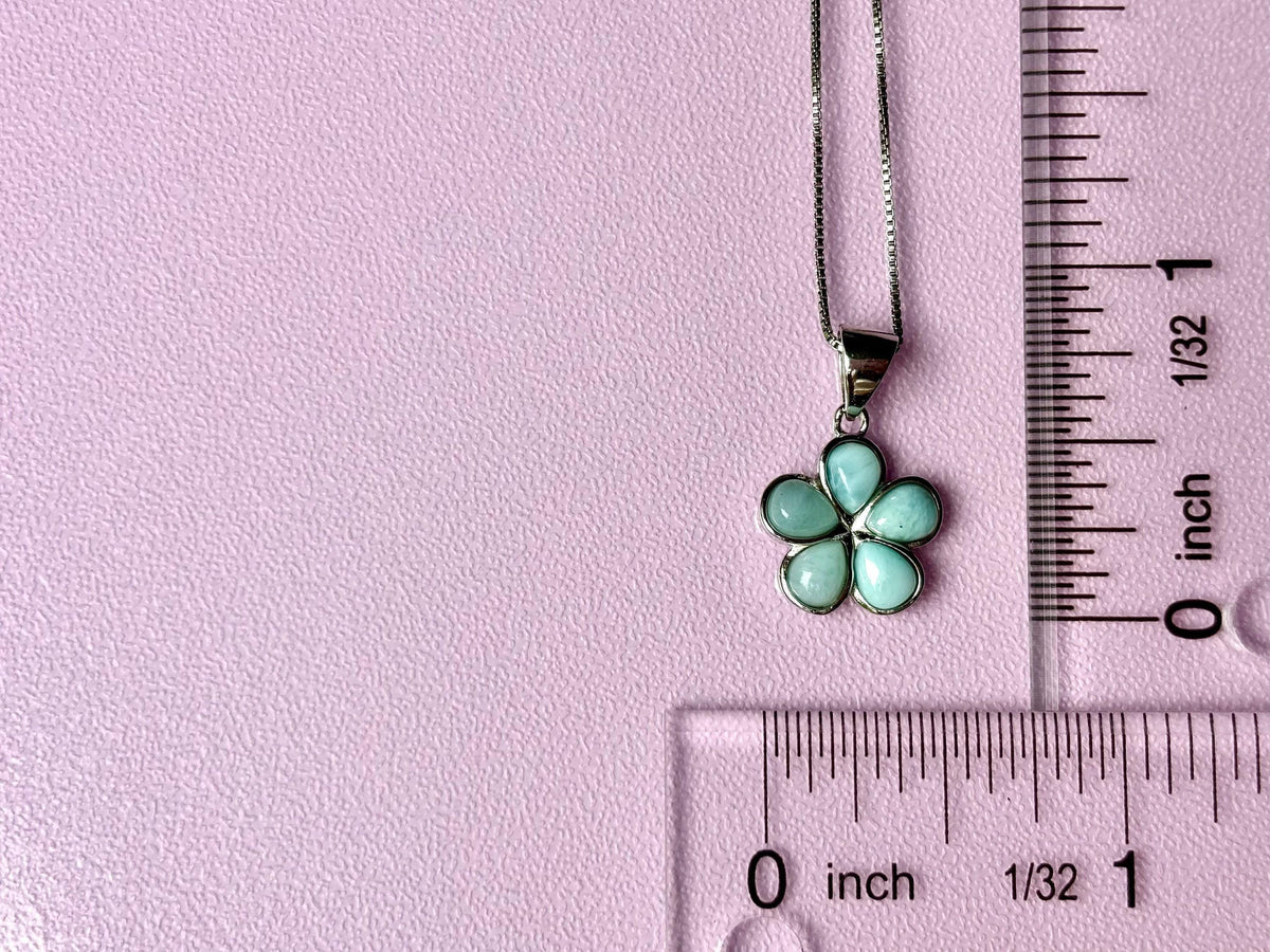 A necklace with a flower pendant is placed on a pink surface, with a ruler for scale.
