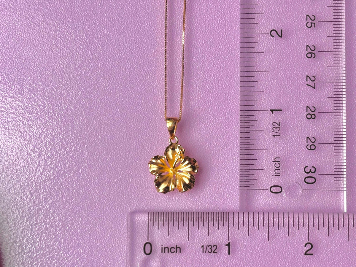 A gold-colored pendant necklace with a flower-shaped pendant is placed next to a ruler on a pink background.