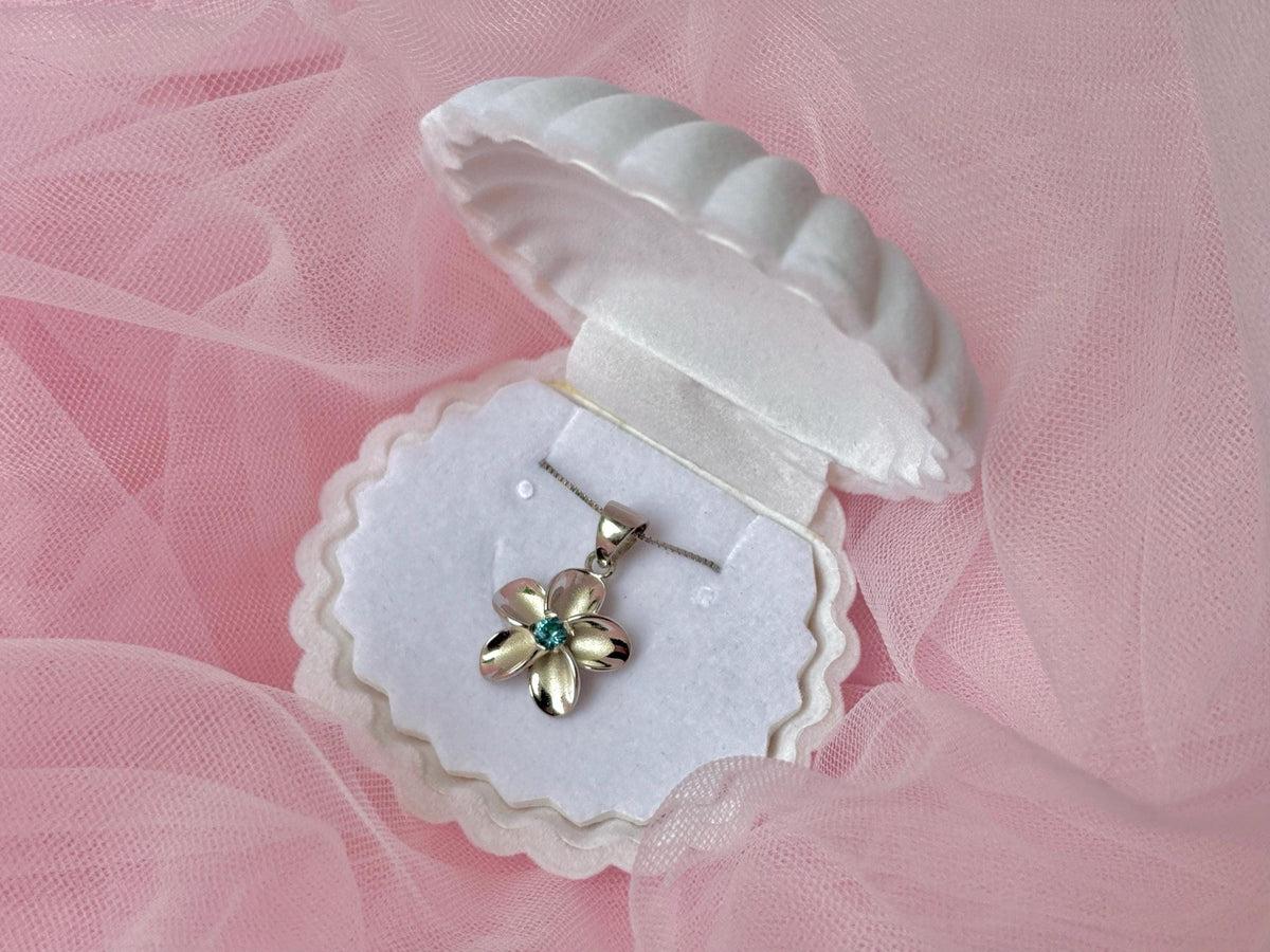A silver necklace with a flower pendant is displayed in a white jewelry box on a pink background.