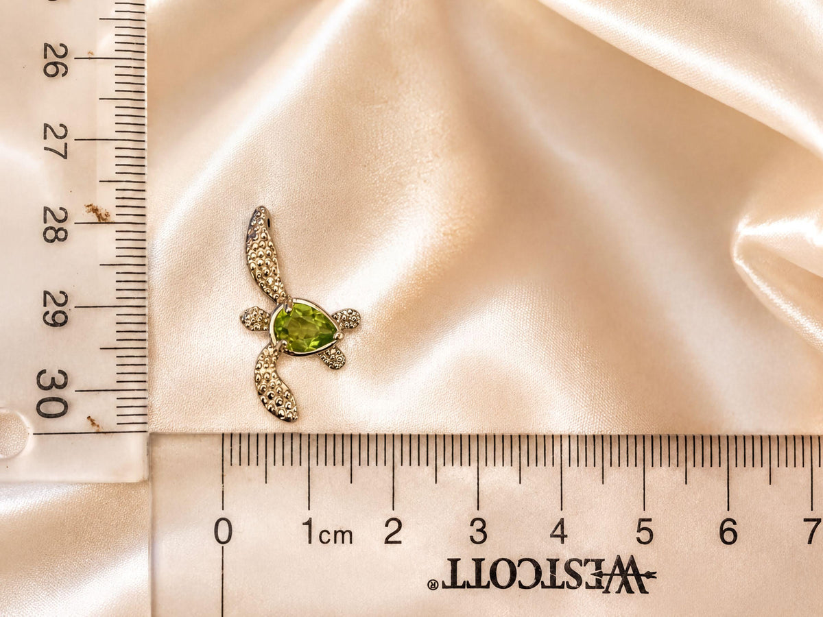 A close-up image of a small turtle-shaped pendant with a green gemstone, placed on a ruler for size comparison.