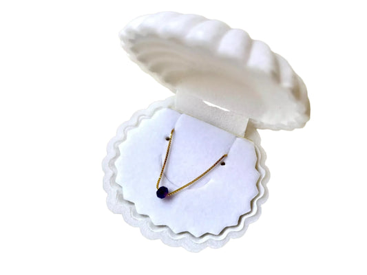A necklace with a blue gemstone is displayed in a white, scalloped-edge box, which is open and revealing the necklace.