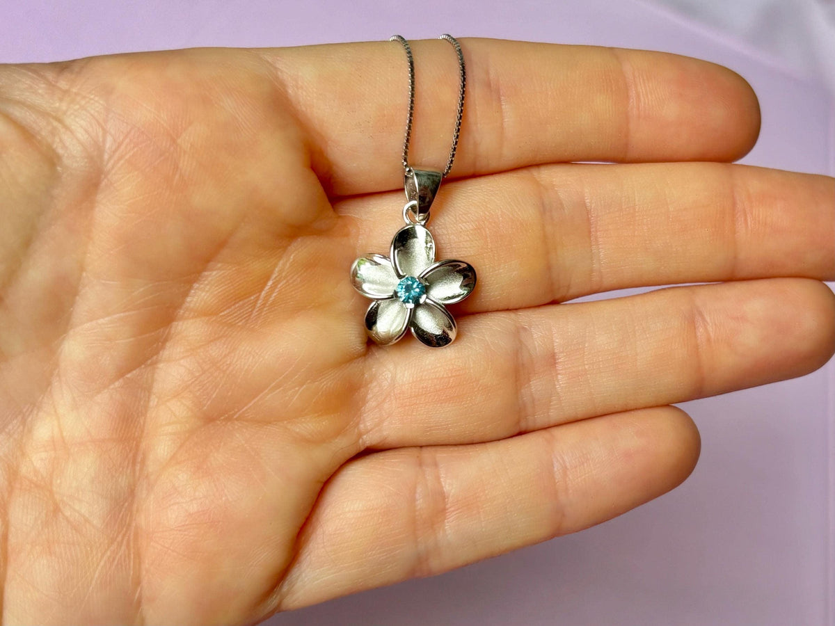 A hand holding a silver flower pendant with a blue gemstone.