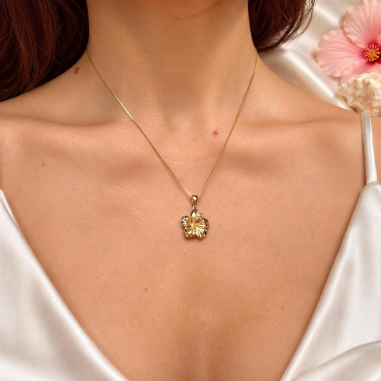 A close-up of a woman wearing a gold necklace with a pendant, and a pink flower is visible in the background.