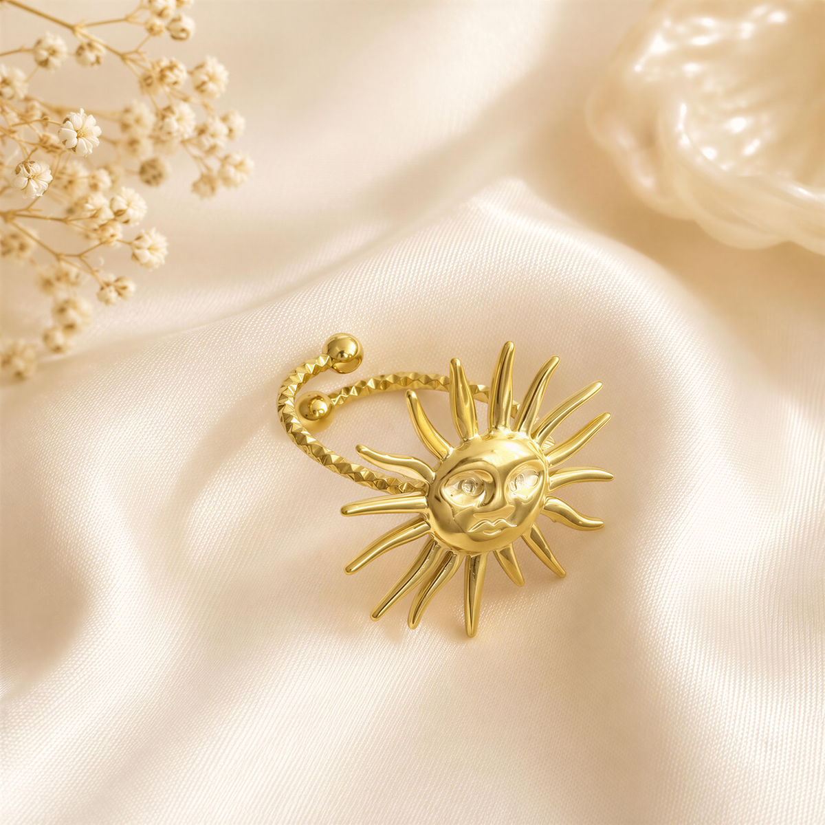 A gold ring with a sun-shaped design is placed on a white fabric surface, accompanied by delicate white flowers.