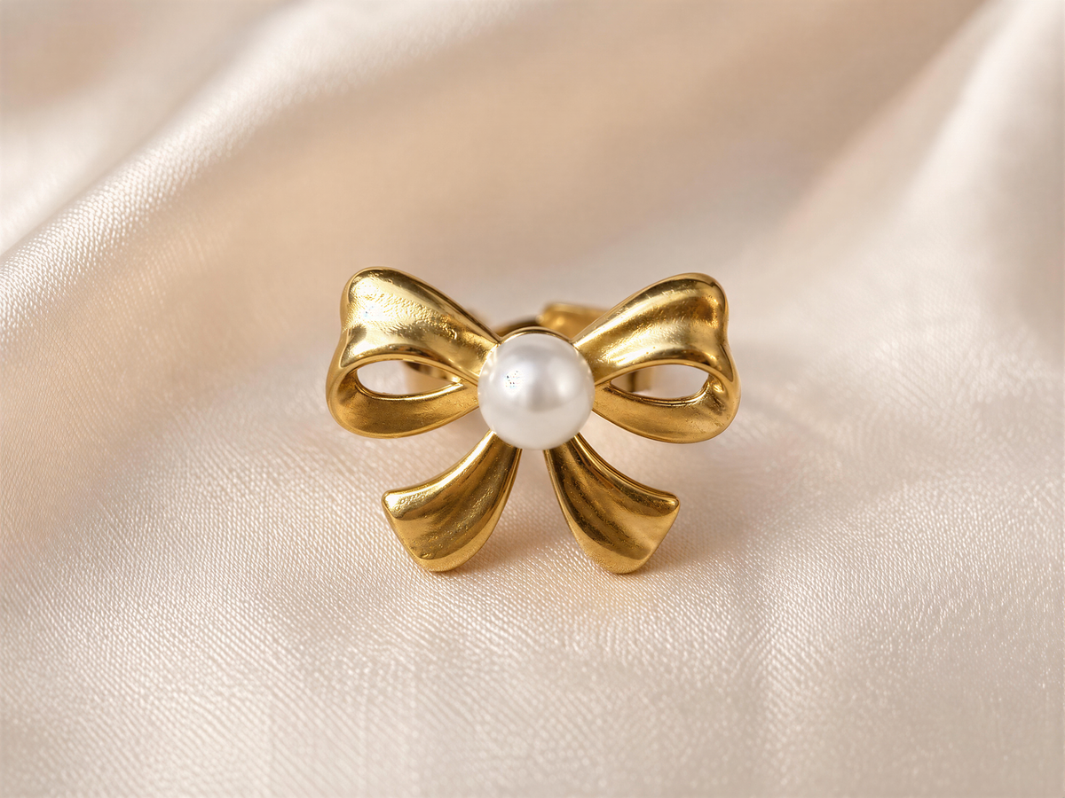 A gold-colored bow ring with a white pearl at the center, placed on a white fabric background.
