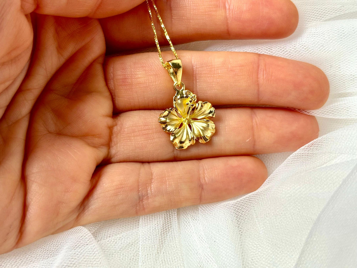 A gold-colored pendant necklace with a small flower-shaped pendant is being held in a person's hand.