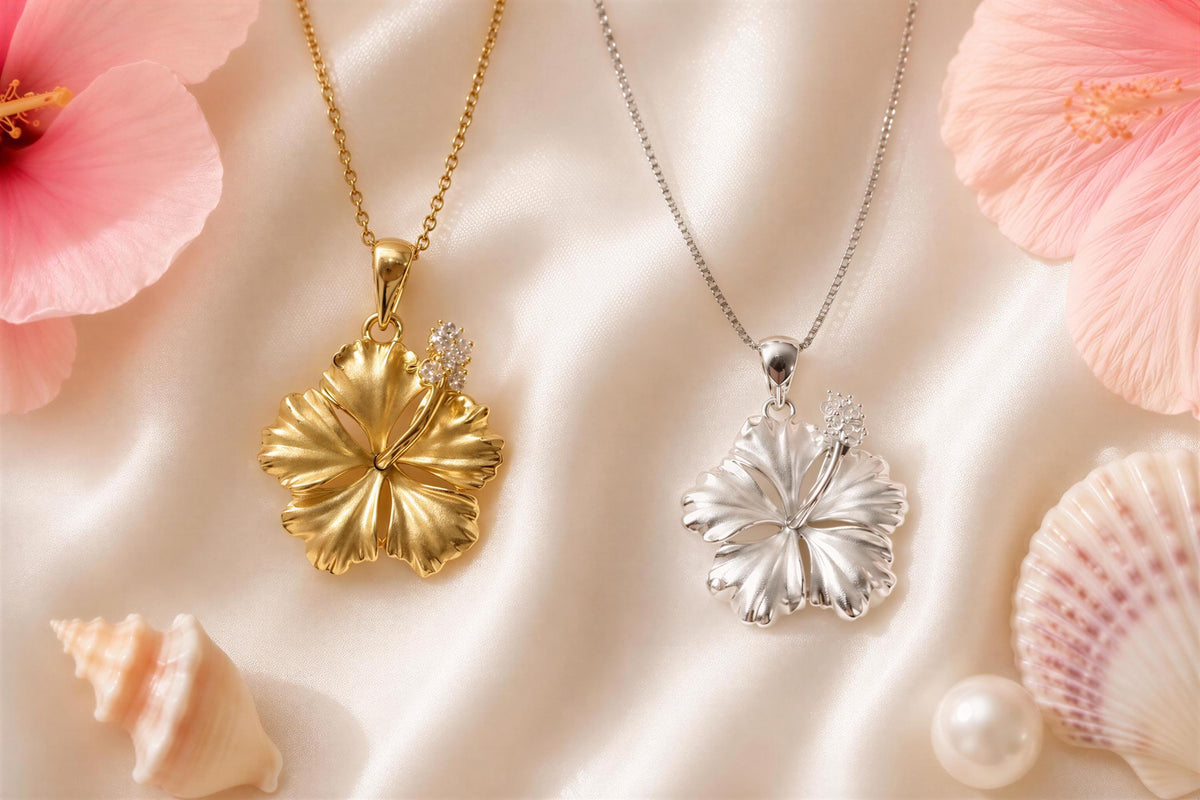 two pendant necklaces with flower-shaped pendants, one in gold and the other in silver, placed on a white background with pink hibiscus flowers and seashells.