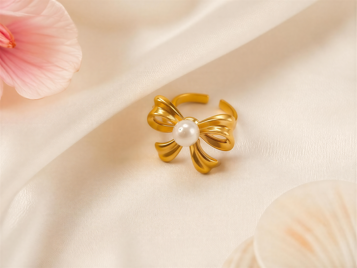 A gold ring with a pearl at the center, placed on a white fabric surface, with a pink flower and seashells in the background.