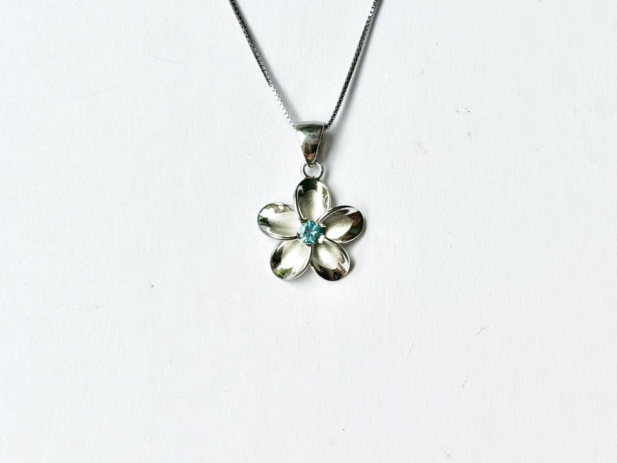 A silver flower pendant with a blue gemstone is displayed against a white background.