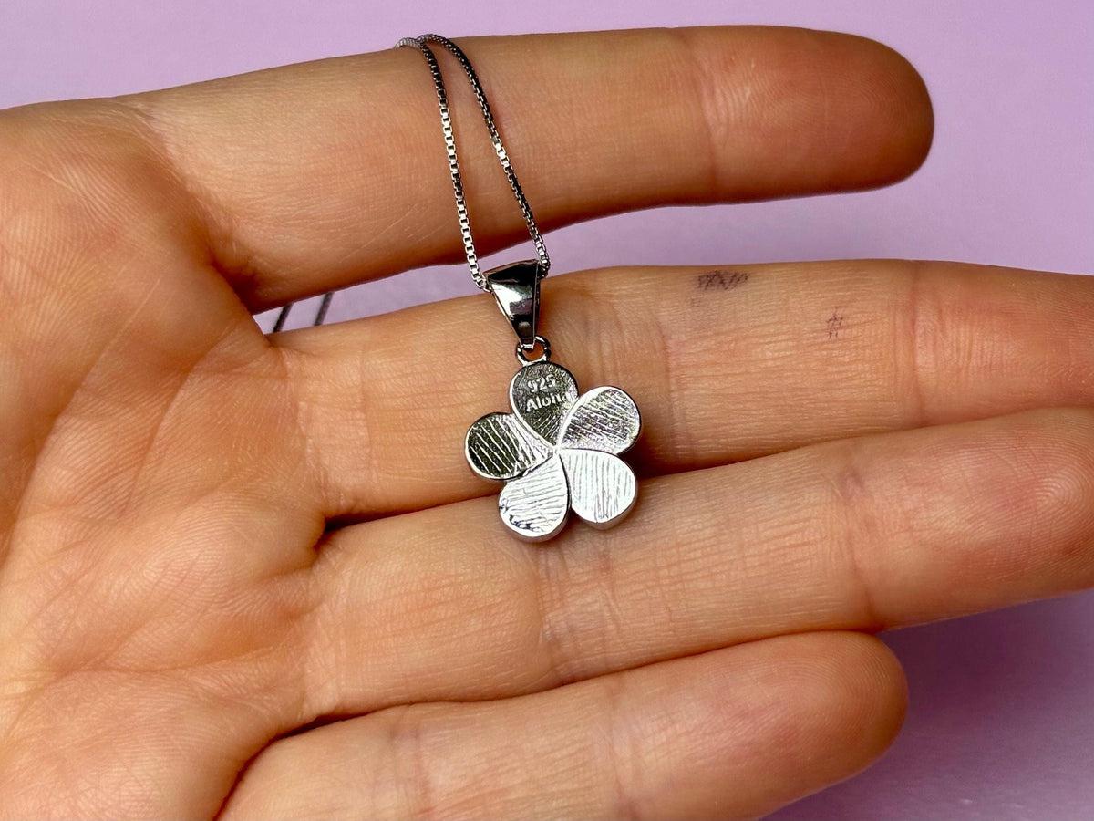 A silver flower pendant necklace is being held in a person's hand against a purple background.